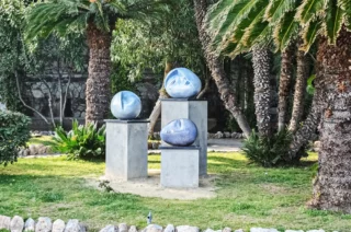 tre sculture di ylli plaka in gres, all'interno di un giardinetto circondato da palme sulla passeggiata di celle ligure. tre sfere ceramiche azzurre, con mezzi volti scolpiti nella ceramica
