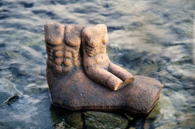 scultura in gres dell'artista e scultore ylli plaka raffigurante due busti senza testa. uno ha un corpo allungato che sembra animale, sta nell'acqua; su di questo sta il secondo busto piÃ¹ piccolo. entrambi in gres